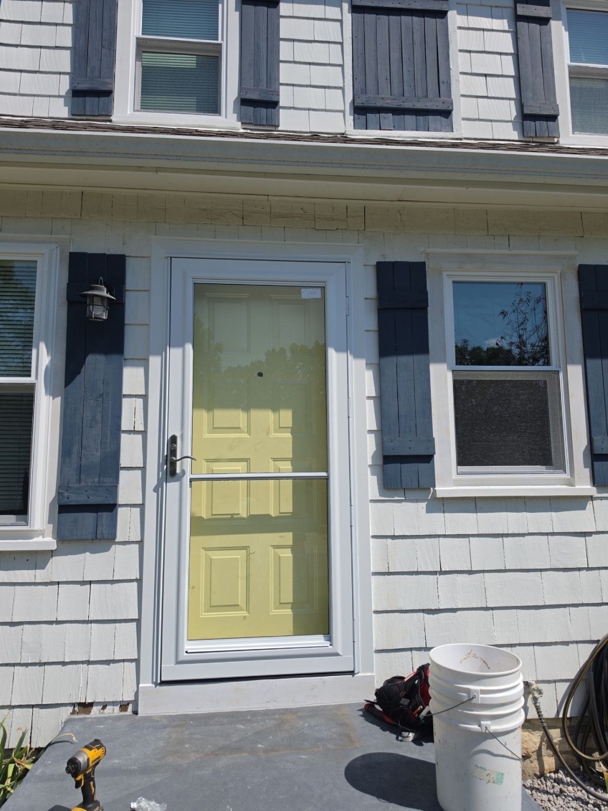 Pale yellow front door with a white storm door and frame, flanked by navy shutters on light shingle siding.