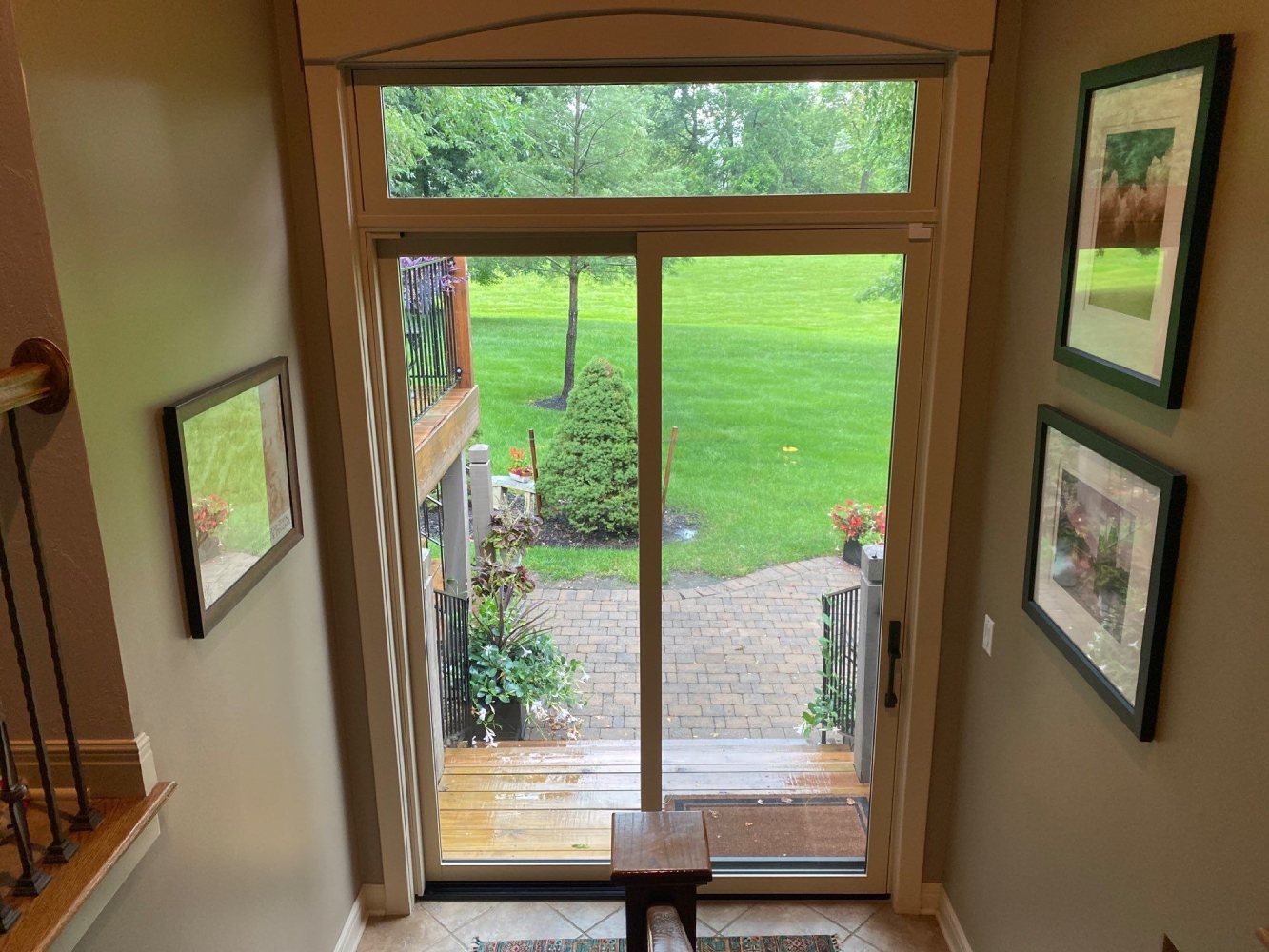 View from inside a home through a wood-framed door with a transom and sliding screen, looking out to a brick patio and green yard.