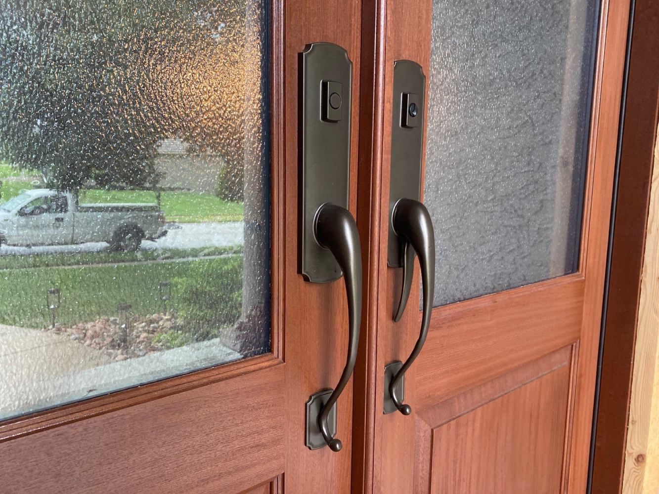 Close-up of a mahogany double door with bronze handlesets and textured glass, reflecting a truck and lawn outside.