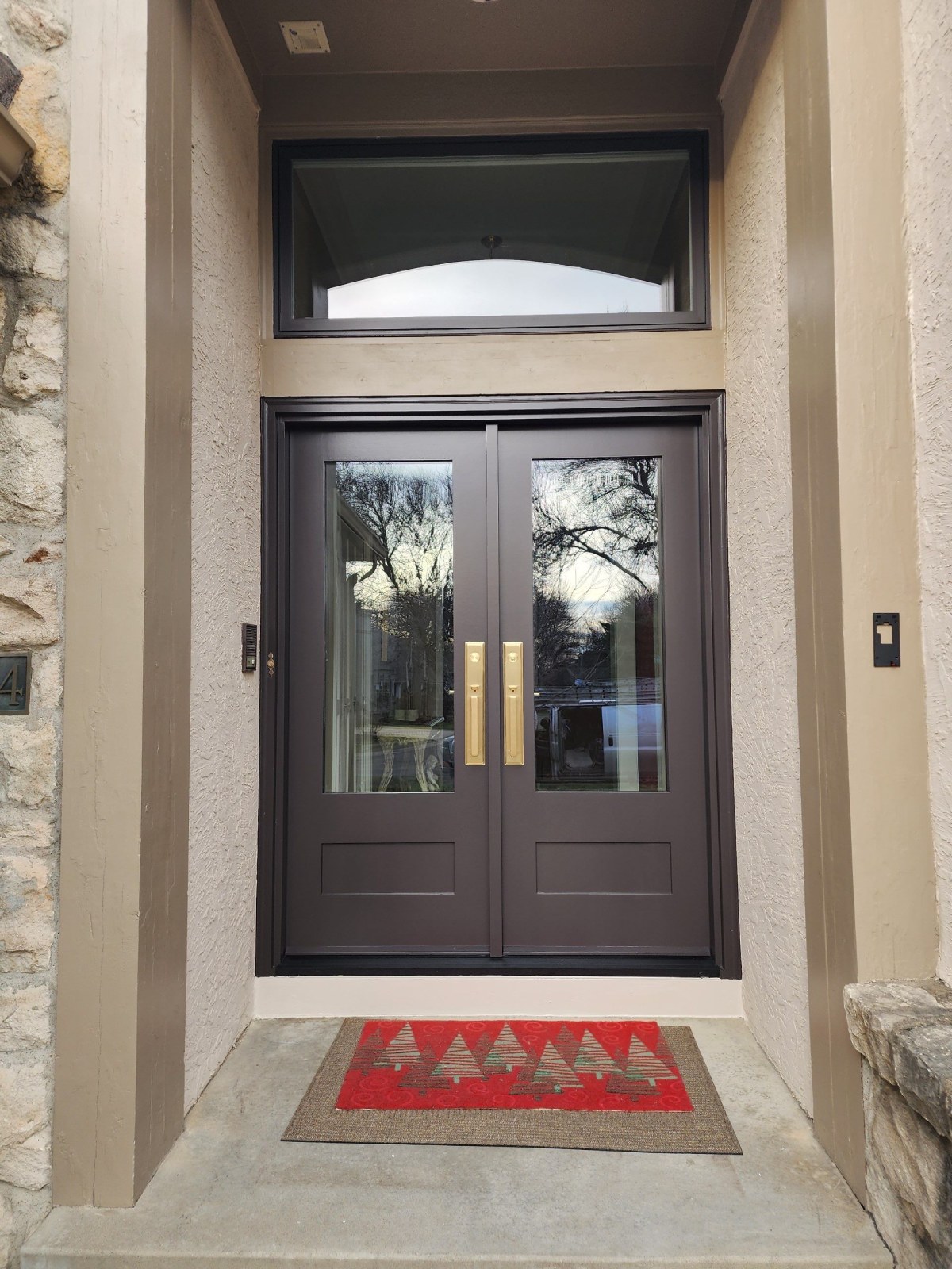 Dark brown double front door with two glass panels and brass handlesets, under a transom in a stucco and stone entry.